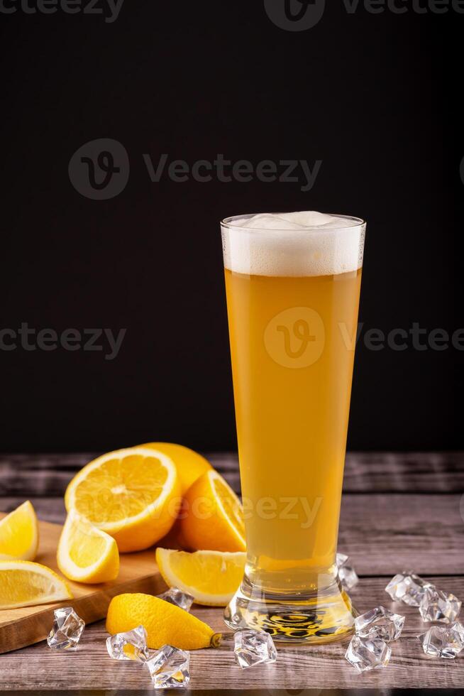 Glass of cold wheat beer with foam beside fresh lemon wedges and ice cubes on wooden surface. Concept citrus craft beer, summer refreshment and flavor pairing. Cold wheat beer with lemon and ice cubes photo