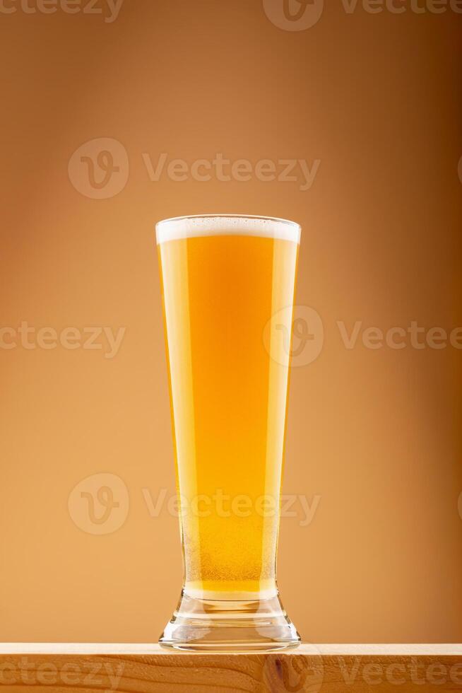 Cold beer with foam served in tall clear glass on wooden surface against warm gradient background. Concept beer clarity, classic lager and refreshing beverage. Cold beer in tall clear glass with foam photo