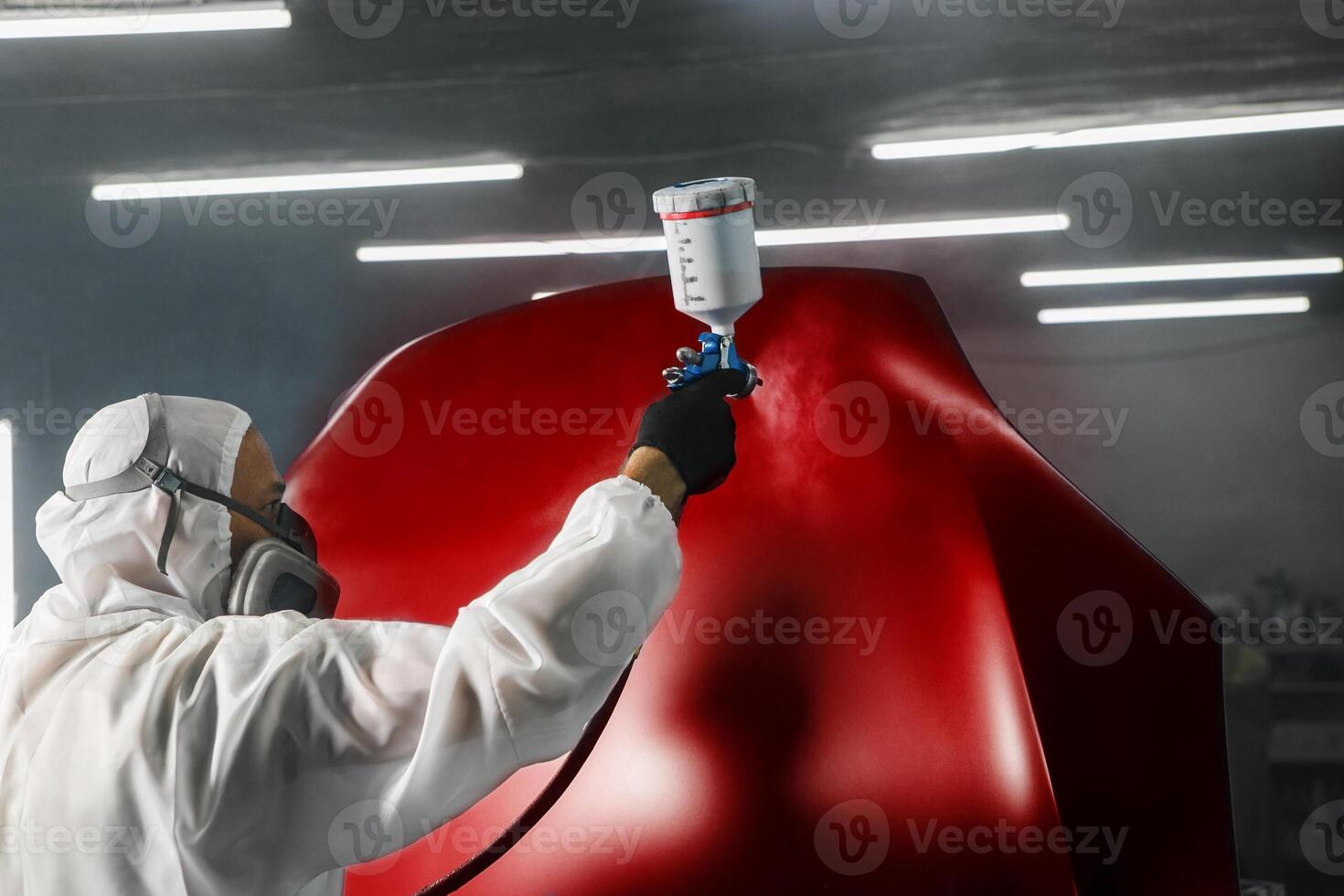 Professional spray painter in protective mask and clothes spraying car hood of vehicle part to red color using spray gun in paint chamber. photo