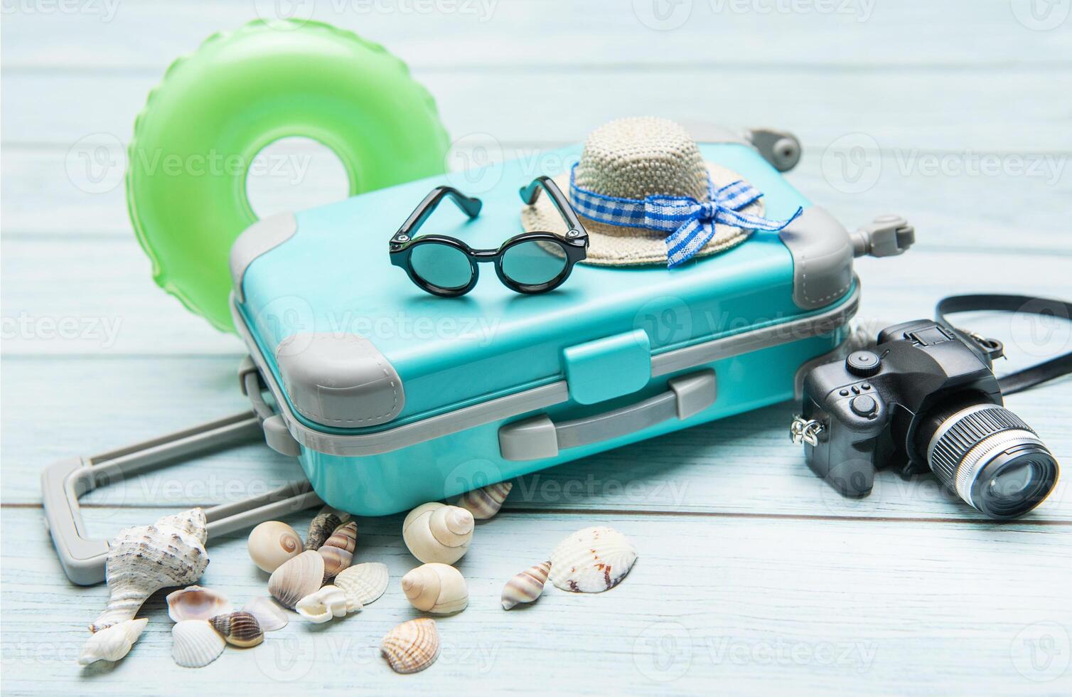 Turquoise suitcase, sunglasses, straw hat, inflatable ring, camera and seashells are laying on light blue wooden surface photo