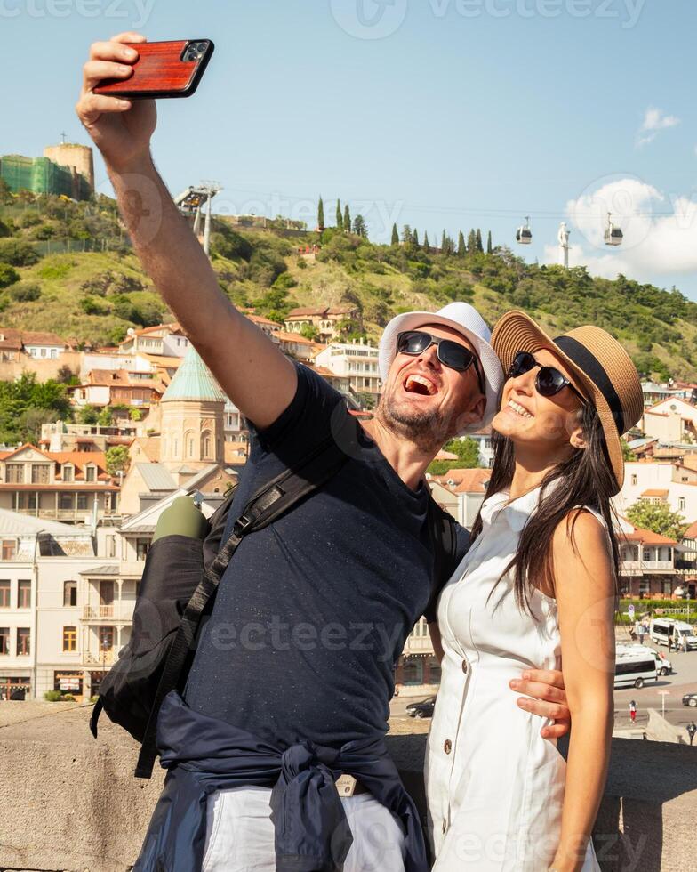 Happy couple takes selfie in front of Tbilisi Old Town skyline with Narikala Fortress and cable car in background, capturing love, connection, travel joy, modern romance. photo