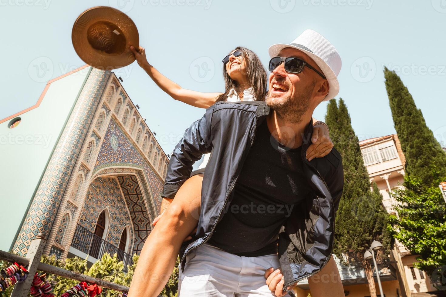 Happy couple enjoying sunny day, man carrying woman piggyback in front of colorful Persian-style mosaic building in Tbilisi old town. Tourist sightseeing famous Georgia attractions in capital Tbilisi photo