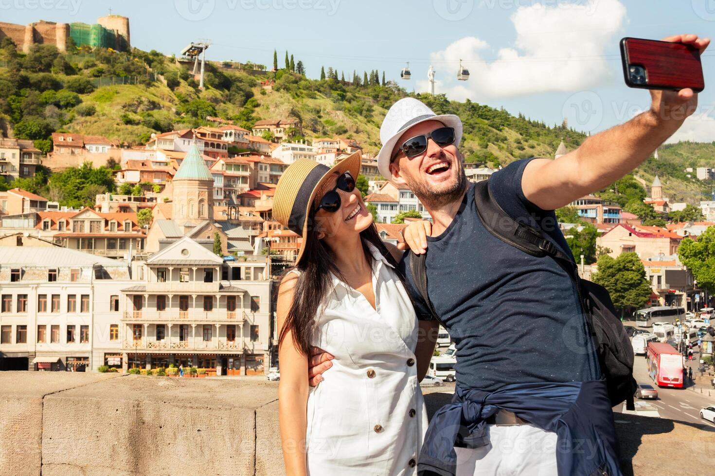 Happy european tourist couple take selfie in Tbilisi old town with Narikala Fortress, Mother Georgia statue, cable cars, and colorful Sololaki hillside houses on sunny summer day photo