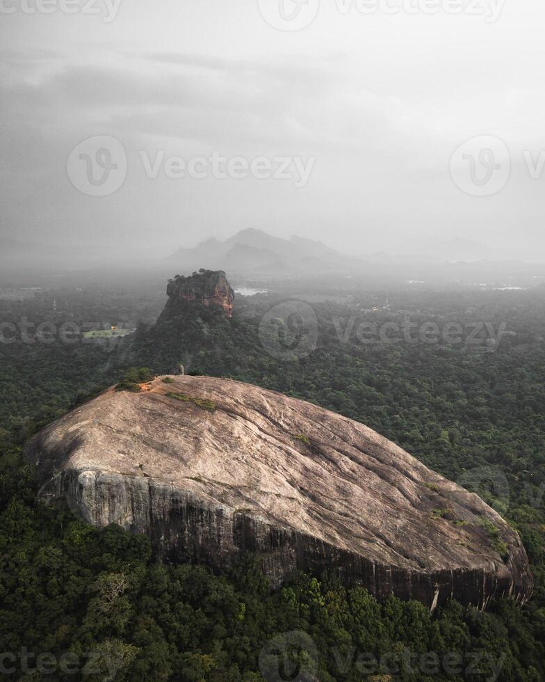 Aerial view Pidurangala Rock with Sigiriya Lion Rock in Central Province Sri Lanka surrounded by dense tropical forest and misty hills under cloudy dramatic sky photo