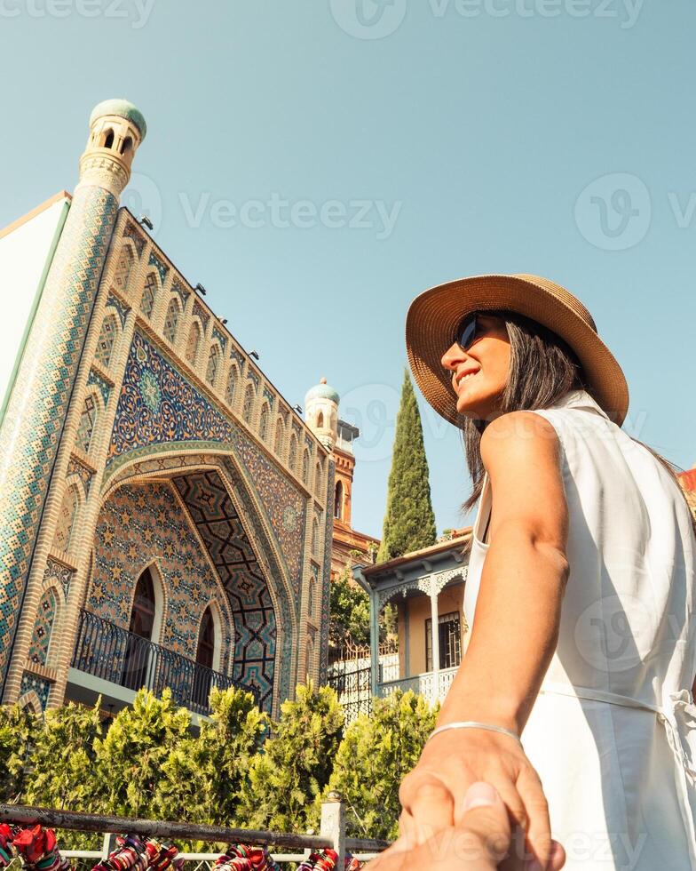 Happy Tourist woman visitor holding partners hand in front of Tbilisi Orbeliani Baths, enjoying colorful architecture in Abanotubani district. Concept travel, connection, trust.Blogger photo hotspot
