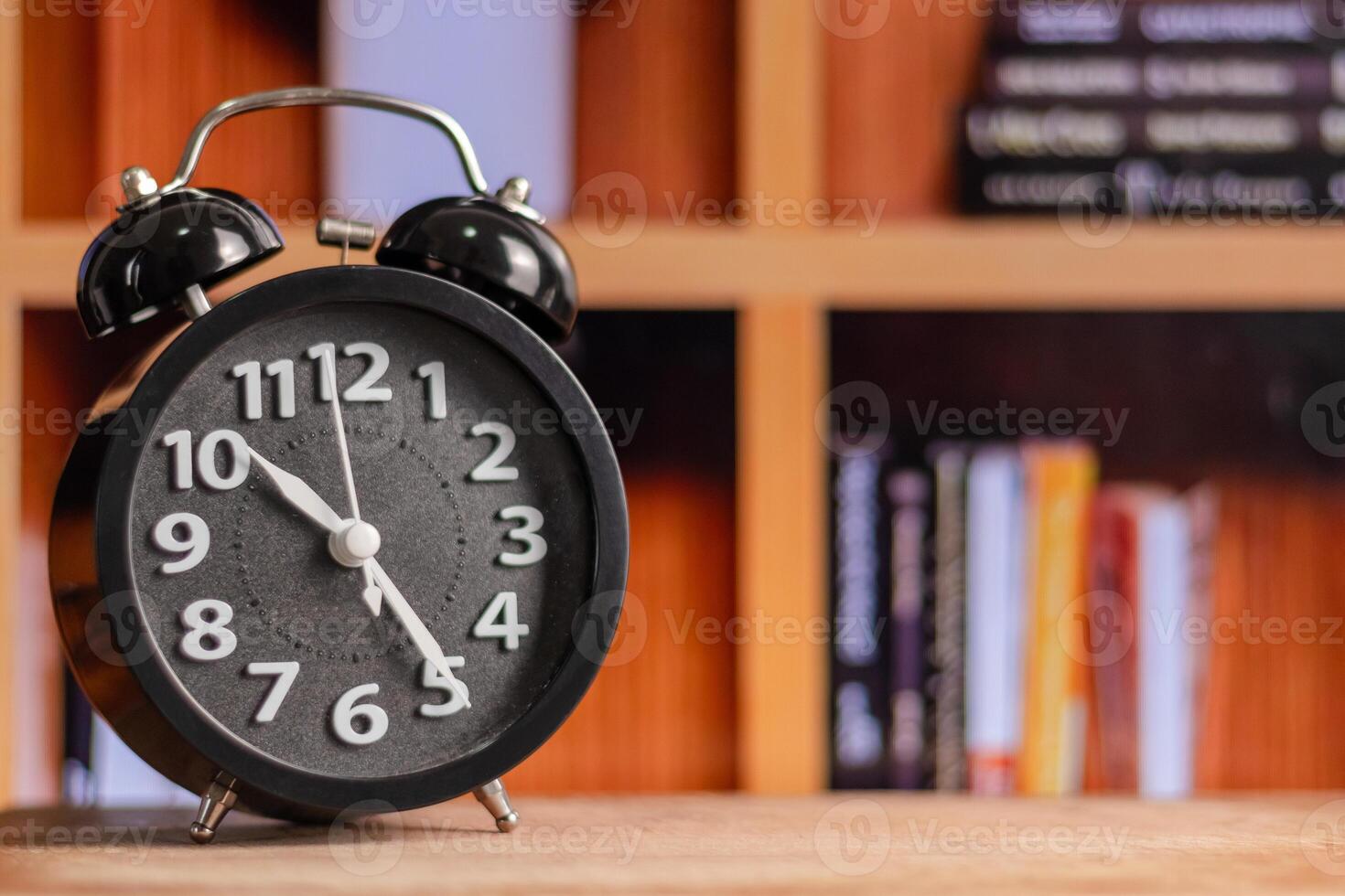 A Black alarm clock on wooden table with bookshelf in the background photo