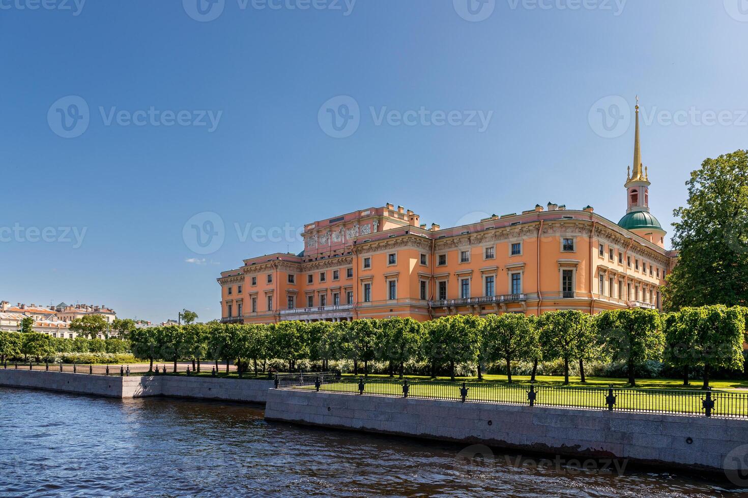 Mikhailovsky Castle with a golden spire stands by the river, with even trees growing along the bank. photo