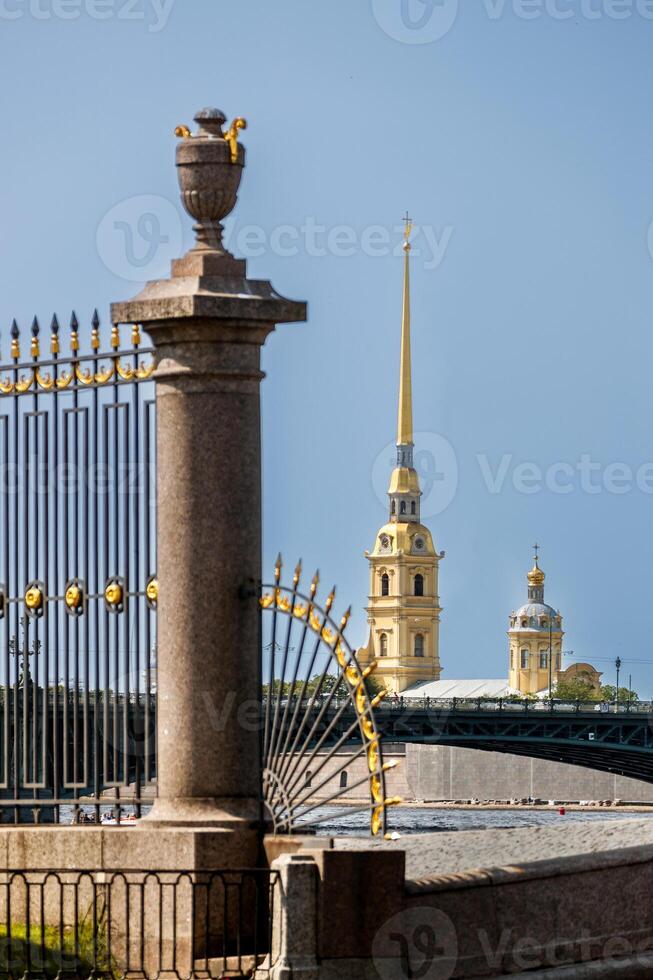 A view of the Peter and Paul Cathedral spire and a bridge over the river, framed by a forged grille and column. photo