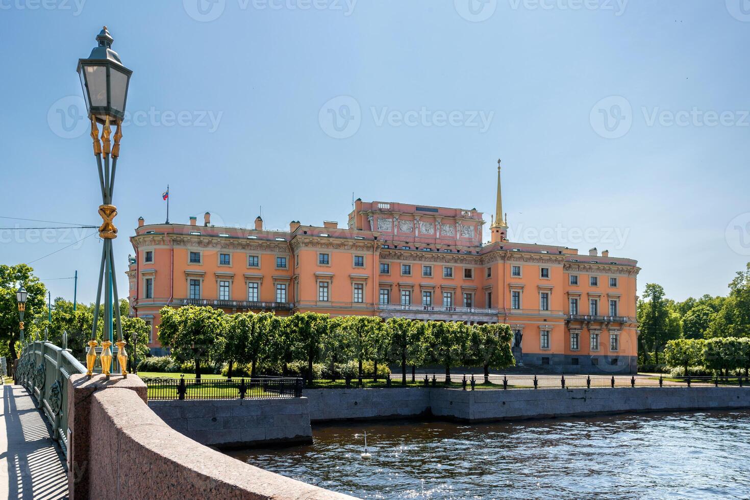 Mikhailovsky Castle, illuminated by the sun, on the river bank, with a bridge and a lantern in the foreground. photo