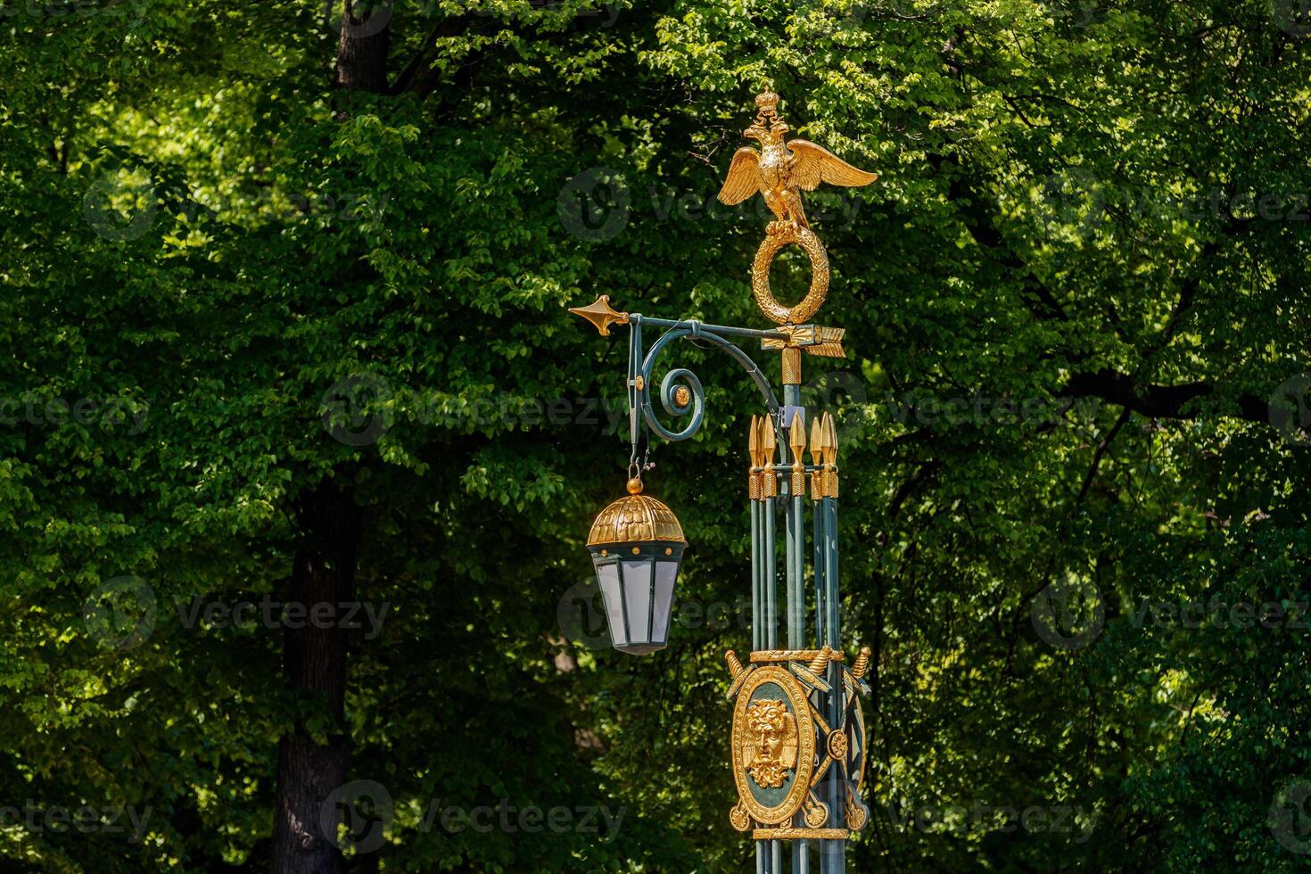A decorative street lamp with a golden double-headed eagle and a lantern, against a background of green tree foliage. photo