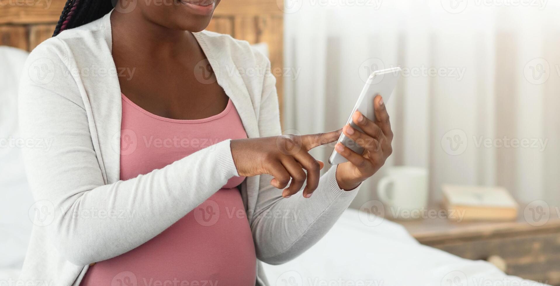 Black Pregnant Woman Doing Online Shopping With Smartphone At Home, Happy Expecting Lady Enjoying Purchasing Goods In Internet, Buying Clothes For Future Baby, Sitting On Bed In Bedroom, Copy Space photo