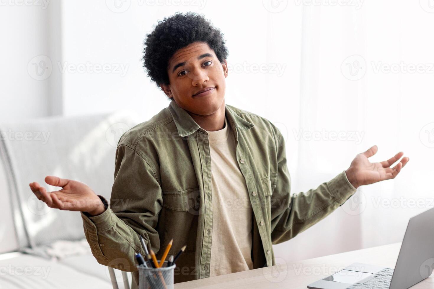 Puzzled black student sitting in front of laptop, not understanding difficult lesson, having difficulty studying online. African American teen guy shrugging shoulders, feeling confused during exam photo