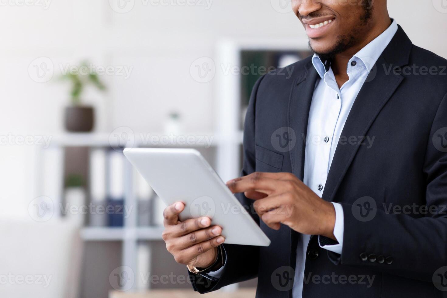 Smiling black manager in suit using digital tablet, office interior, copy space. Happy african american businessman using business application on pad, enjoying user friendly software photo