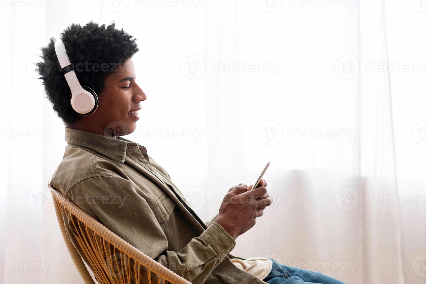 African American teen guy with headphones listening to music on smartphone while sitting in chair at home, copy space. Side view of black teenager relaxing in armchair, enjoying online radio broadcast photo