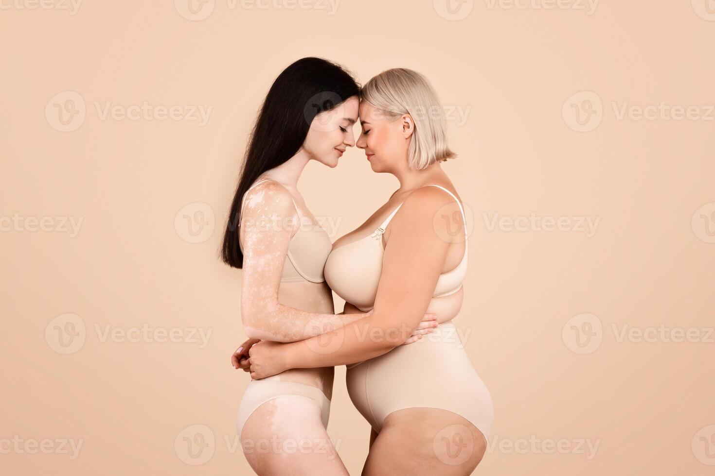 Side View Shot Of Two Body Positive Ladies Standing In Underwear, Young Plus Size Female And Woman With Vitiligo Skin Disorder Embracing And Touching Foreheads On Beige Studio Background, Copy Space photo