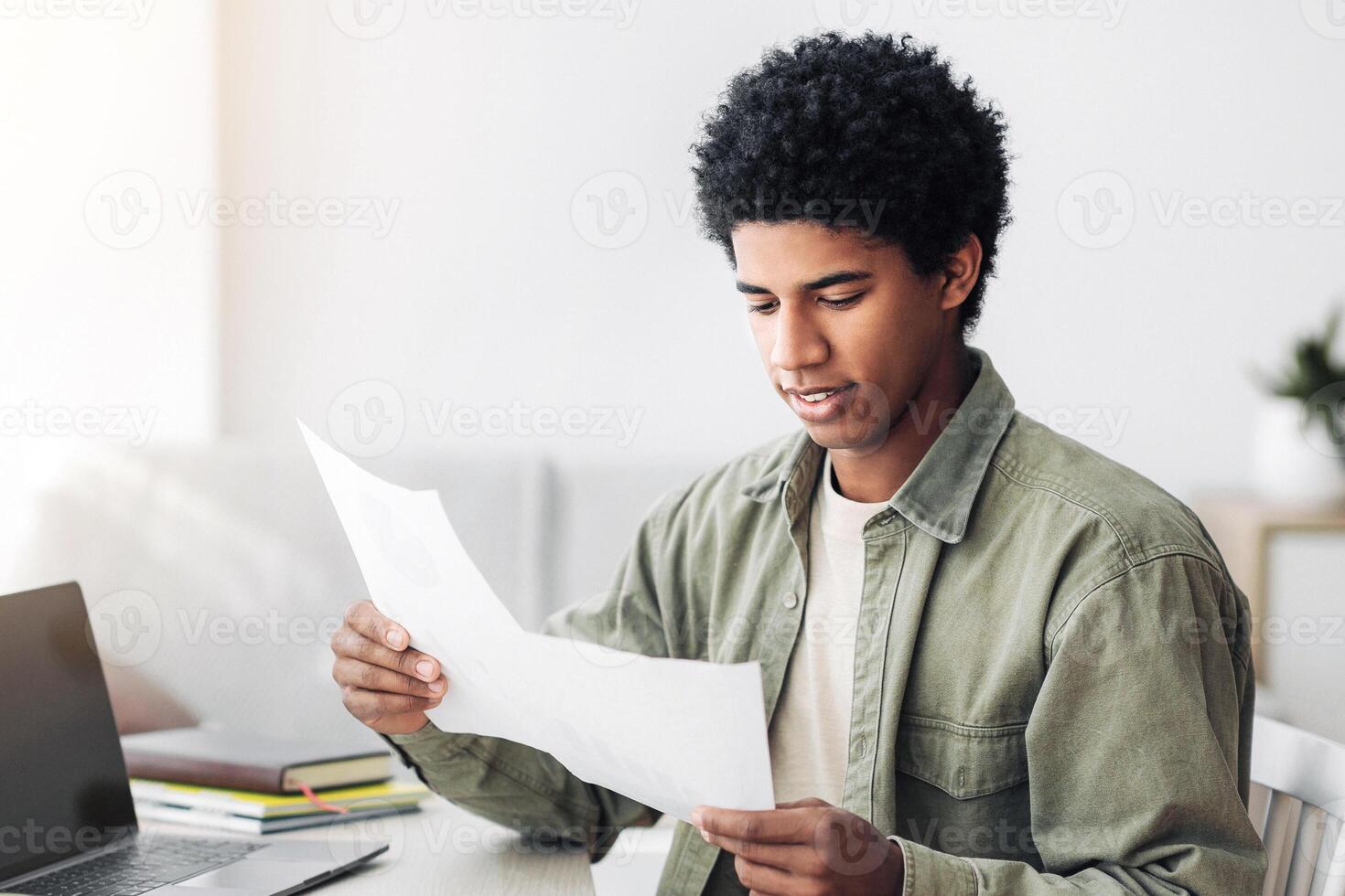Black teenager studying remotely on laptop, working on coursework, coordinating his school project with tutor at home. African American student with papers using PC with empty screen, mockup photo