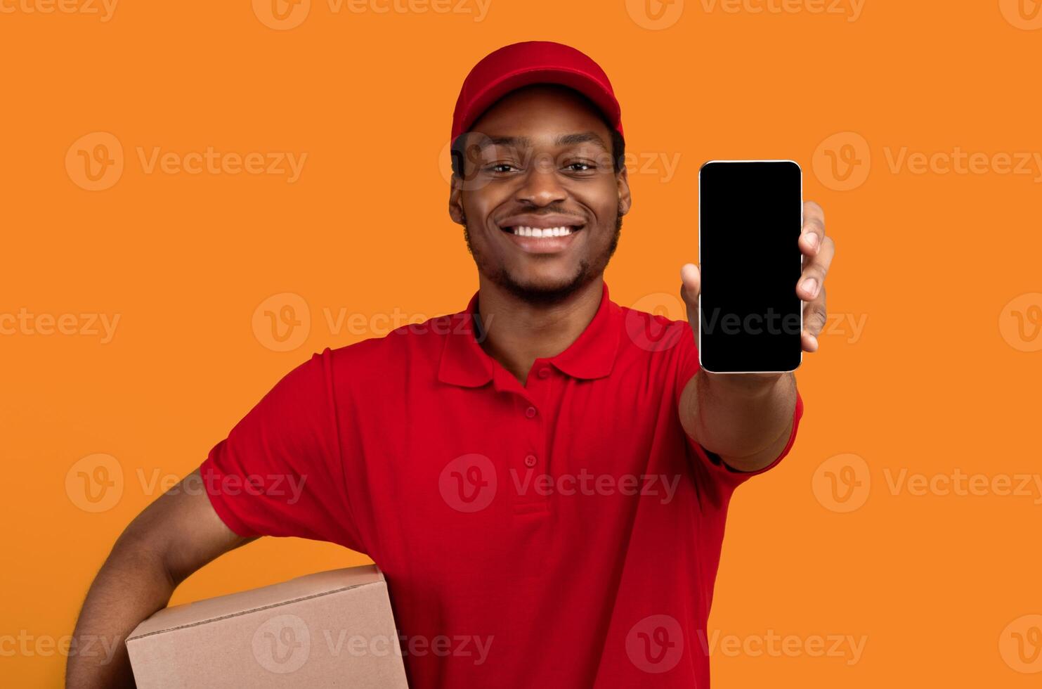 Delivery Service Application. Smiling african american man showing smartphone with blank screen for mockup, wearing red cap, uniform and thermo backpack bag, holding box, isolated on orange background photo