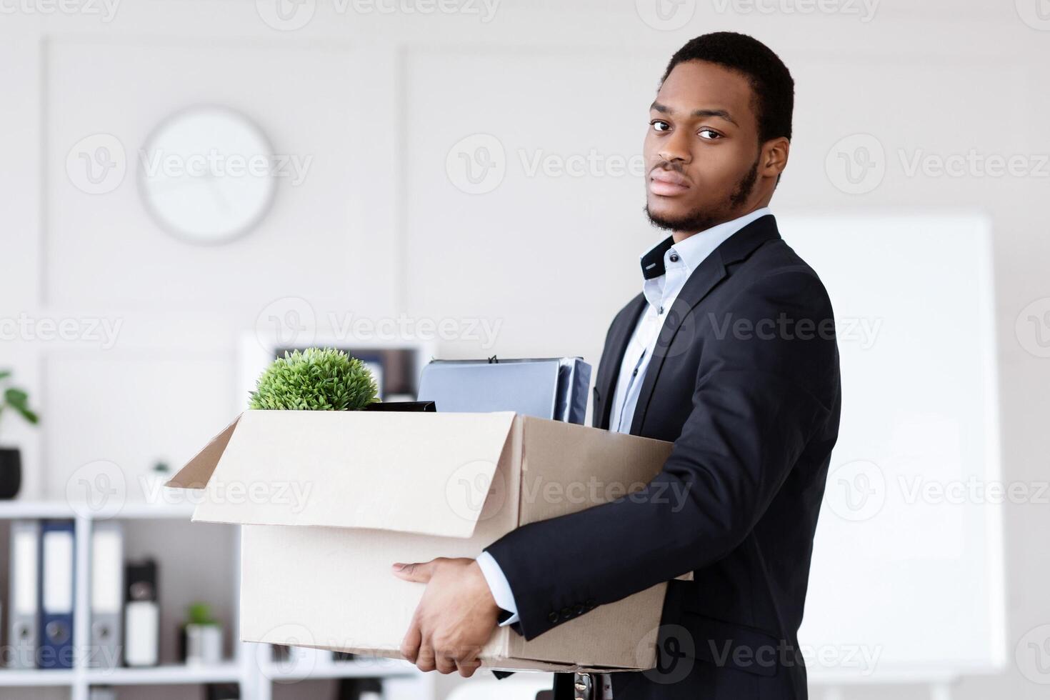 Sad young black man got fired, standing in office and holding box with his belongings, copy space. Terminated african american guy in suit lost his job, packing working stuff before leaving workplace photo