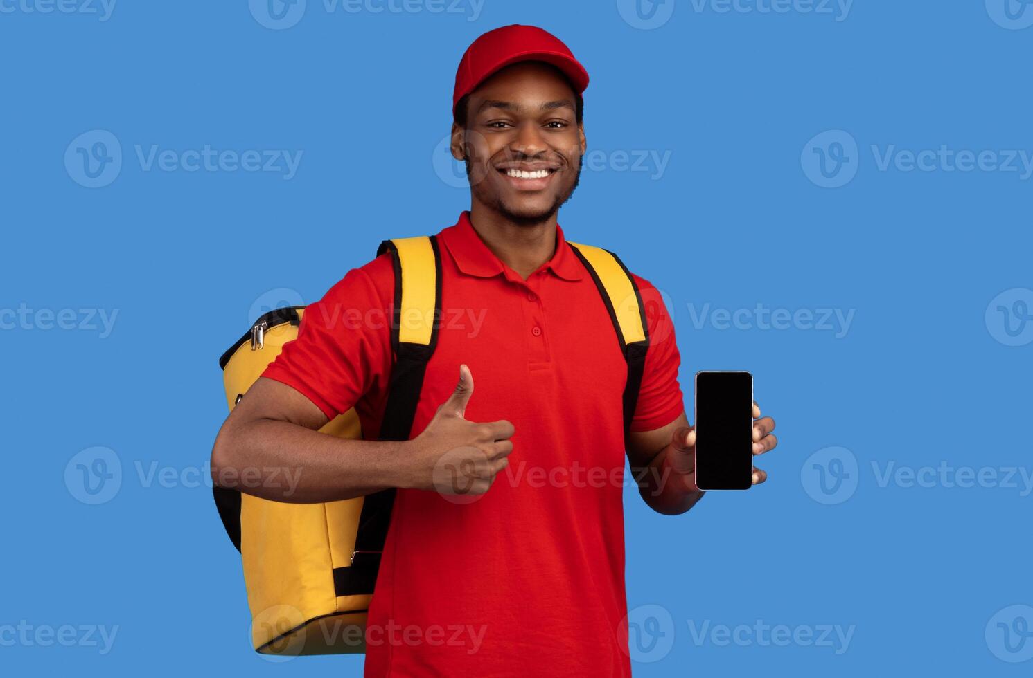 Food Delivery. African american courier man holding mobile phone with blank screen for mockup, wearing red cap, uniform and thermo backpack bag, showing thumbs up isolated on blue background photo
