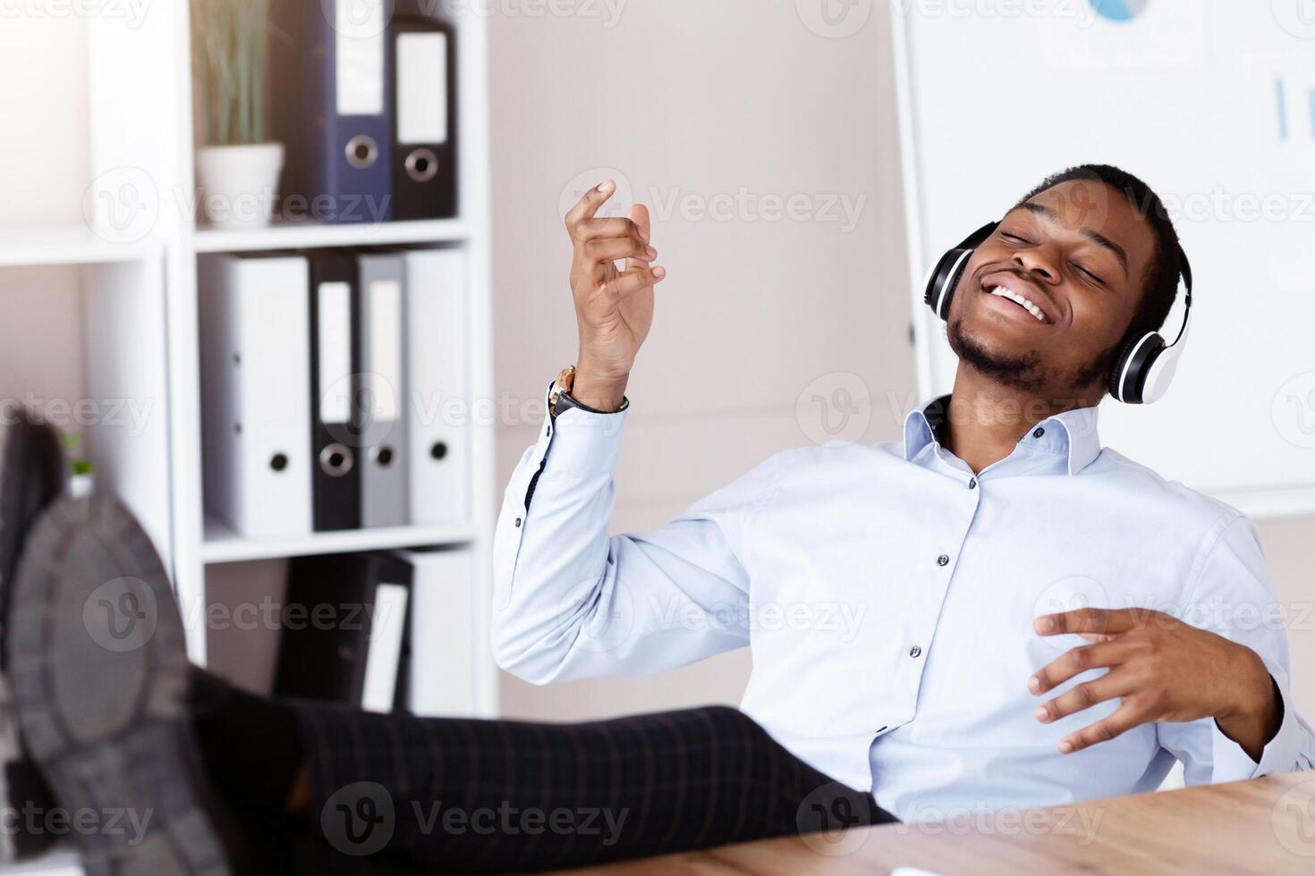 Stress relief concept. Relaxed african american young man businessman with closed eyes listening to music during working day in office, playing invisible guitar, using wireless headset, empty space photo