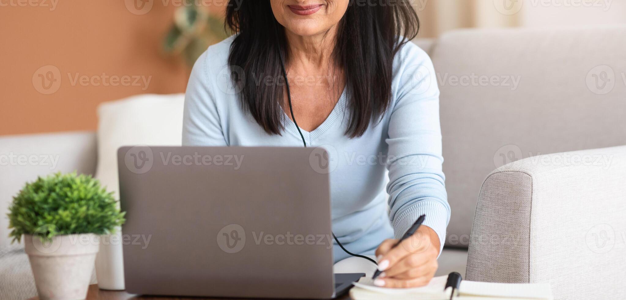 Modern mature smiling woman in headset taking online course or training on computer at home. Smart positive caucasian female in headphones watching webinar making notes studying distant on laptop photo