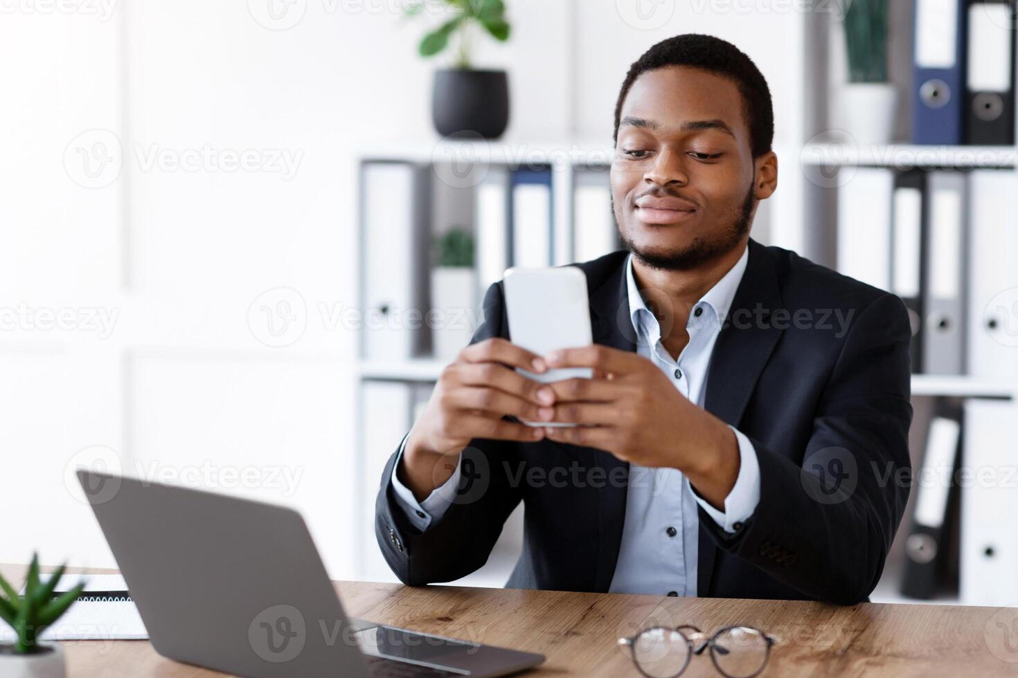 Young african american man entrepreneur in suit using smartphone in office, sitting at workdesk with laptop and cup of coffee, chatting with clients or checking emails on mobile phone, copy space photo