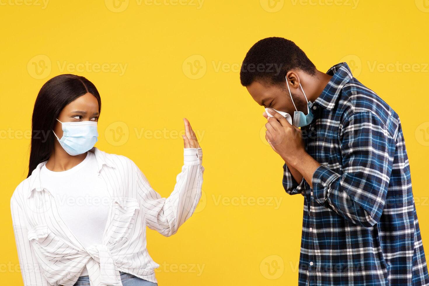 Health care and covid-19 pandemic. Sad african american lady in protective mask shows stop sign to man, guy sneezes and has first symptoms of disease, isolated on yellow background, studio shot photo