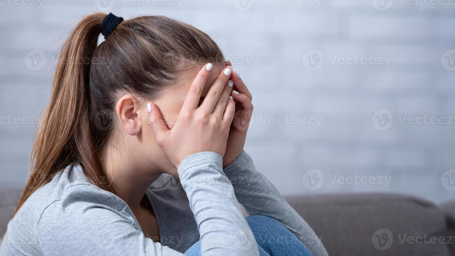 Side view of young woman with depression covering her face and crying on sofa at home. Upset millennial lady suffering from mental disorder, having psychological problem, feeling stressed and lonely photo