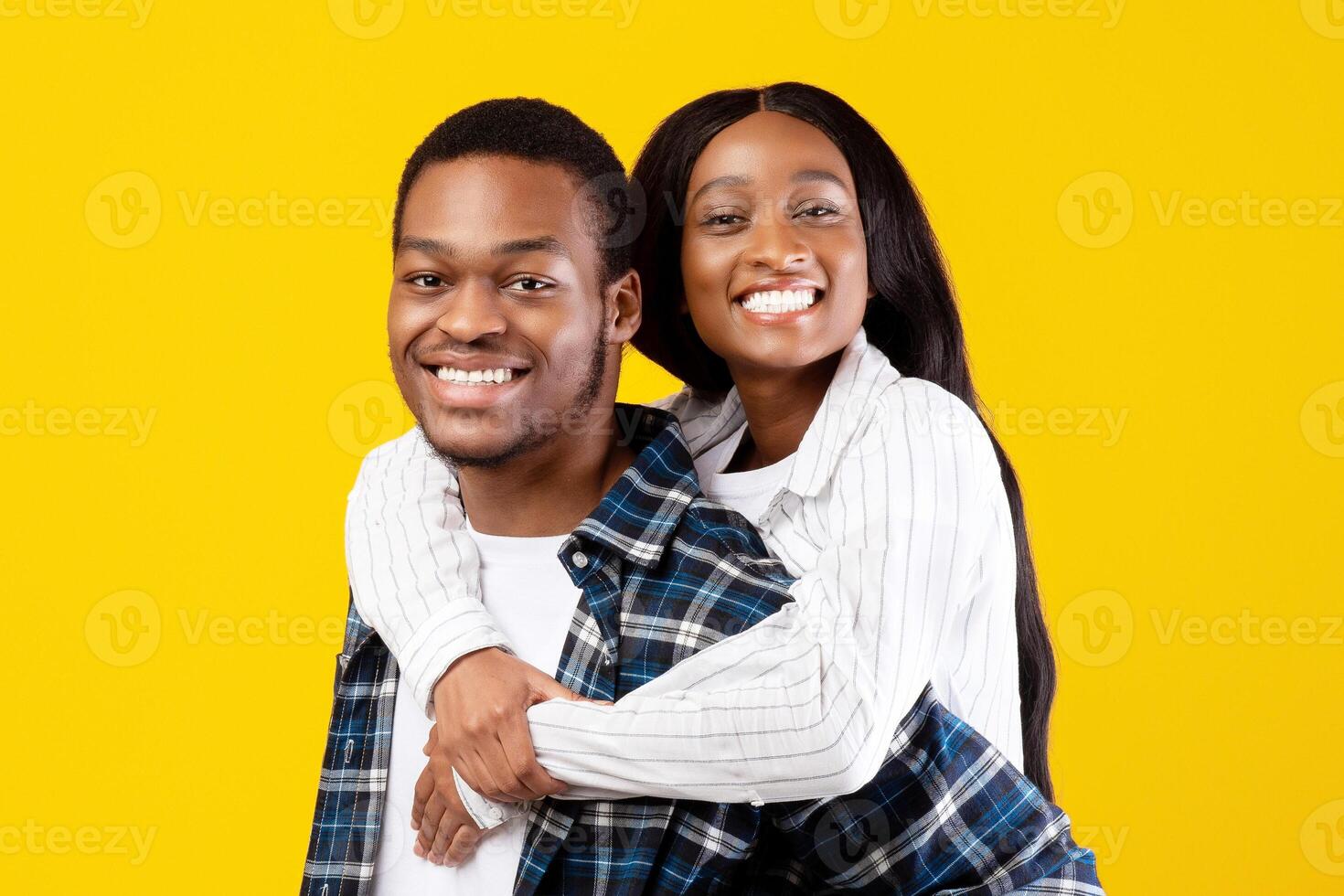 Happy man carrying girlfriend on his back, fun together and enjoying date. Smiling young african american guy holding lady and look at camera isolated on yellow background, studio shot, free space photo
