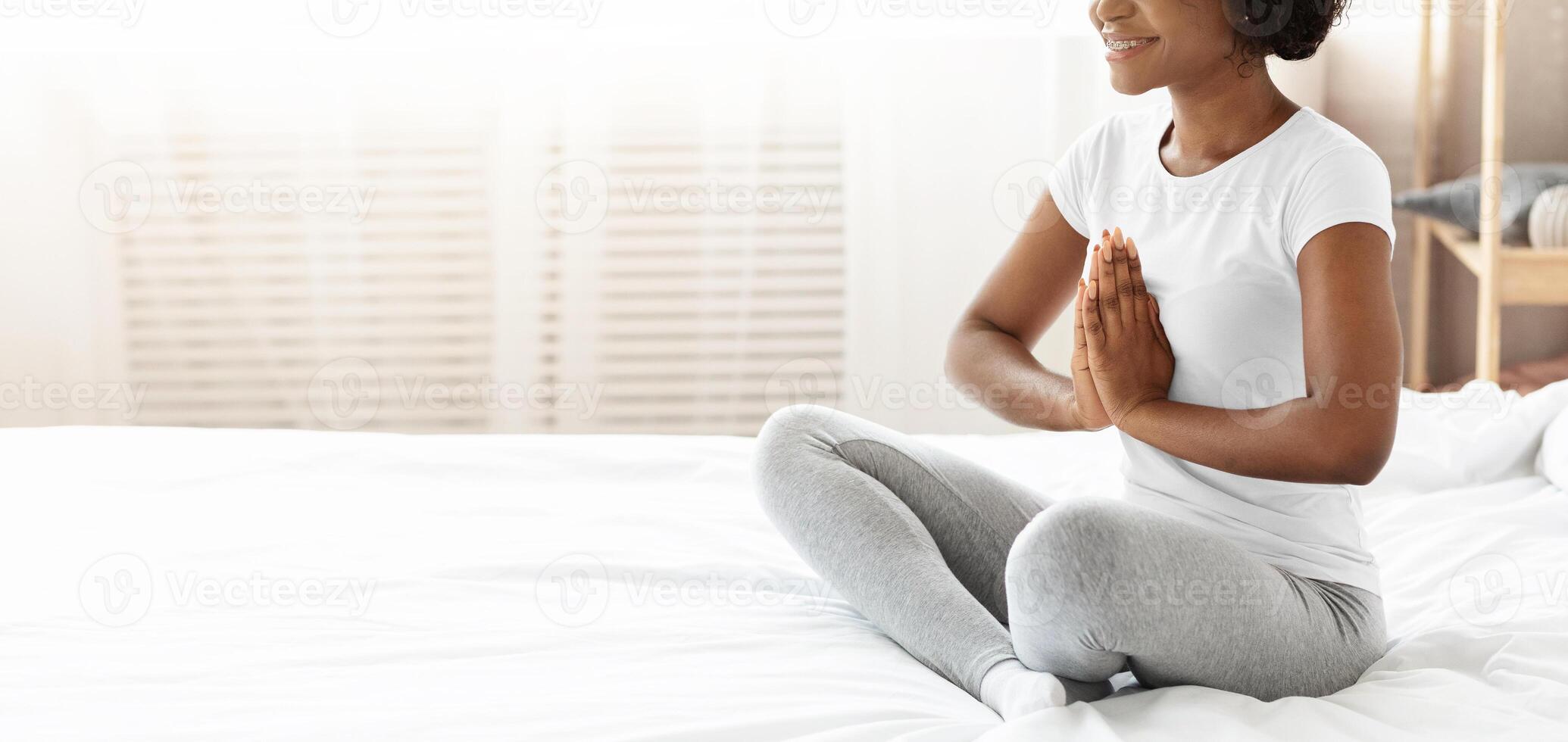 Cropped of black woman in pajamas practicing yoga at home, panorama with copy space. Unrecognizable african american lady sitting in lotus pose on bed, starting her morning with exercising photo