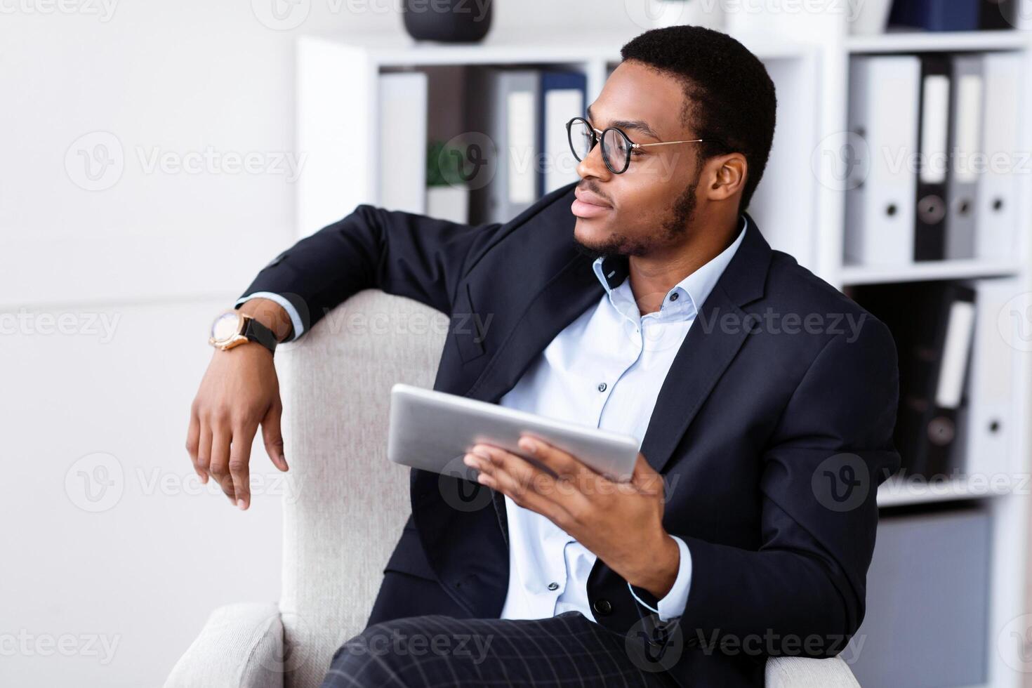 Thoughtful black businessman in stylish suit and glasses sitting in chair in office, using digital tablet while having break, looking at copy space. African american entrepreneur looking for new ideas photo