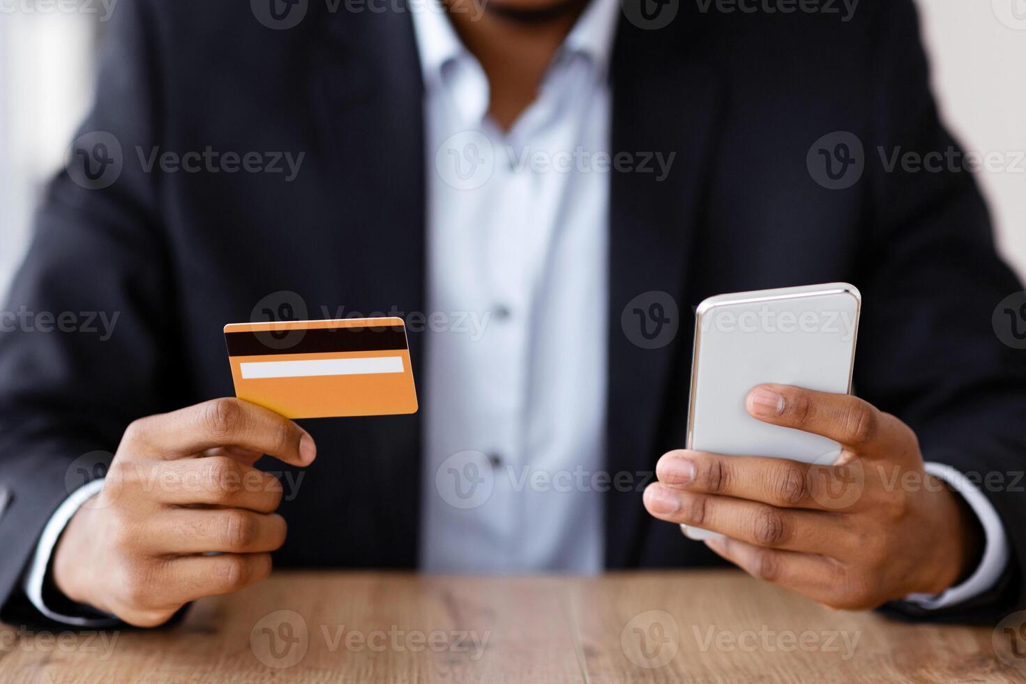 Unrecognizable black man with mobile phone and credit card, shopping online while working in his office, ordering food online for lunch break, african american businessman using e-payment service photo