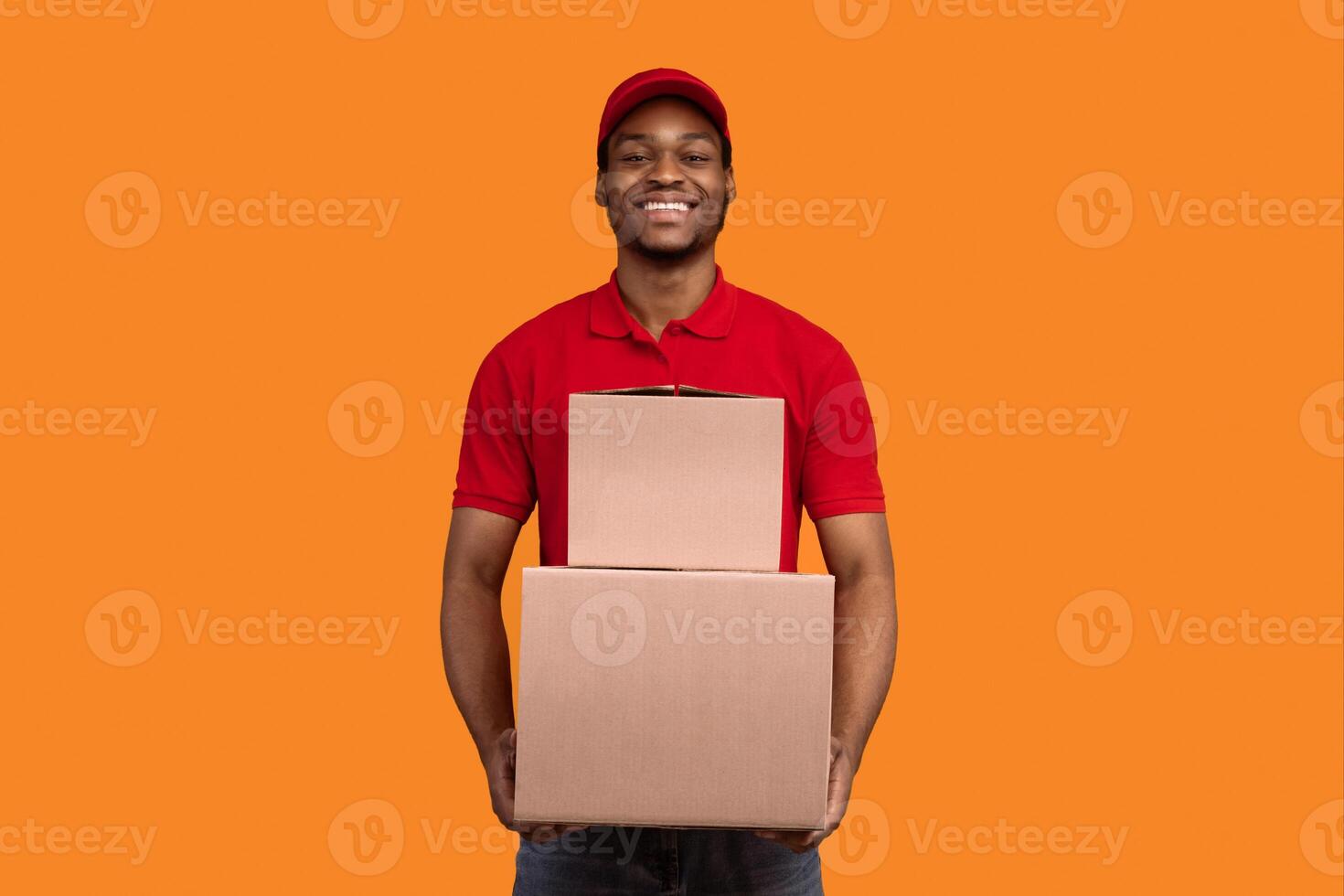 Delivery Service Concept. Smiling african american male courier in red cap and uniform holding pile of cardboard boxes, carrying packages, standing isolated over orange studio background photo