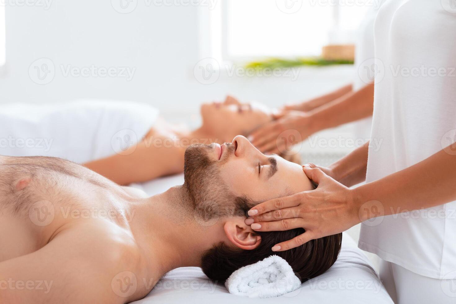 Spouses Receiving Head Massage Lying With Eyes Closed Relaxing On Beds At Spa Indoors, Focus On Husband. Body Relaxation And Pampering, Wellness, Beauty Day For Couple Concept photo