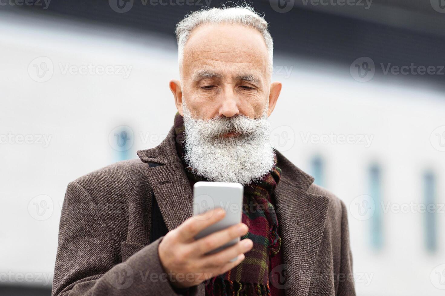 Wealthy senior man checking his modern smartphone while walking by street, panorama with copy space. Handsome grey-haired businessman coming home from job, looking at mobile phone screen photo