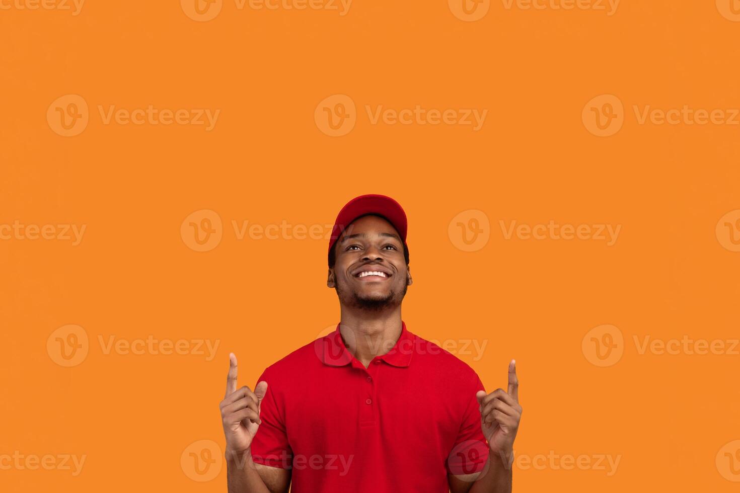 Portrait of optimistic african american delivery man in red t-shirt and cap smiling, looking and and pointing fingers up on copy space text or product isolated over orange studio background photo