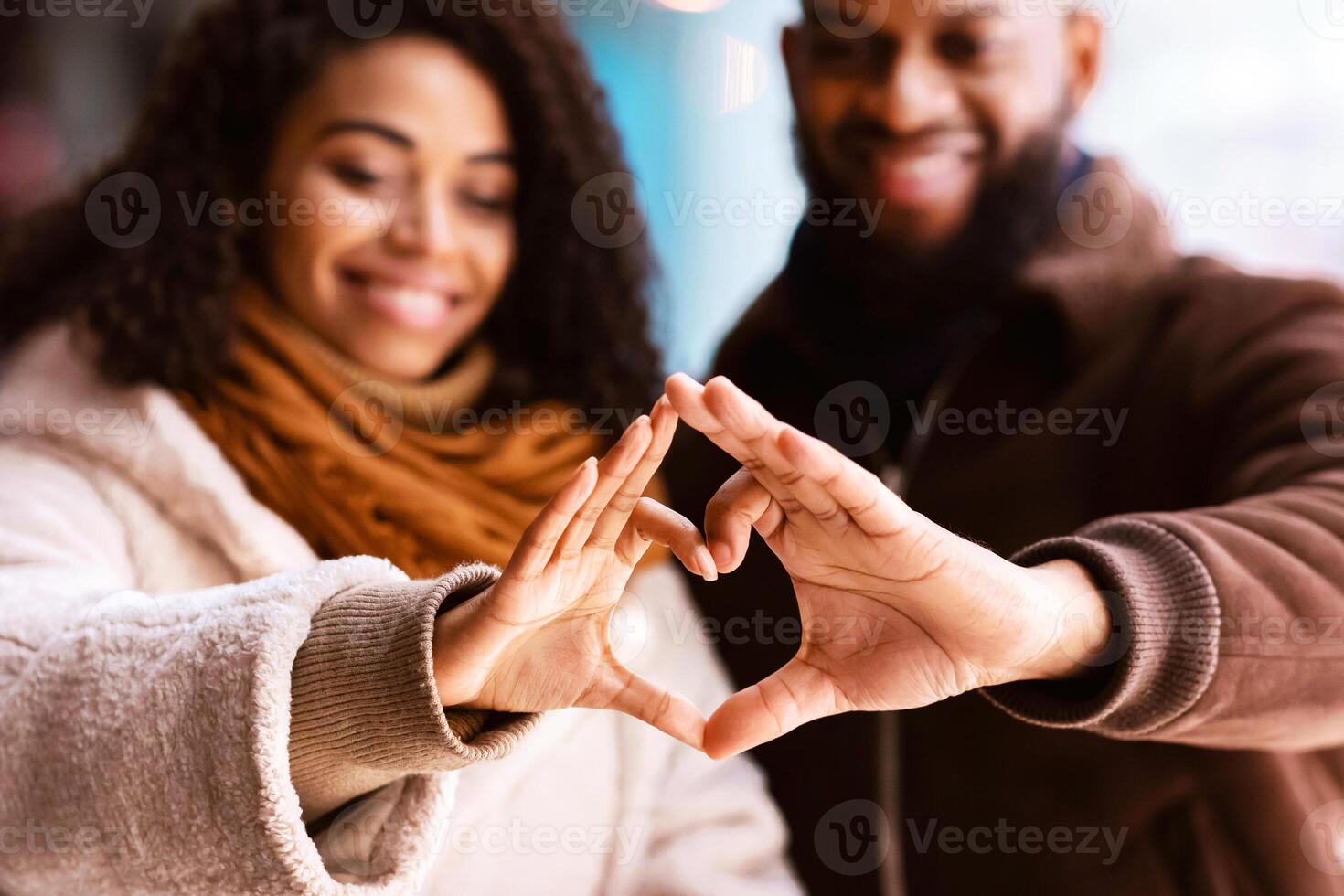 Celebrating Valentine's Day. Close up of smiling loving black man and woman making heart shape with fingers walking together in the evening, looking at hands, selective focus, blurred background photo