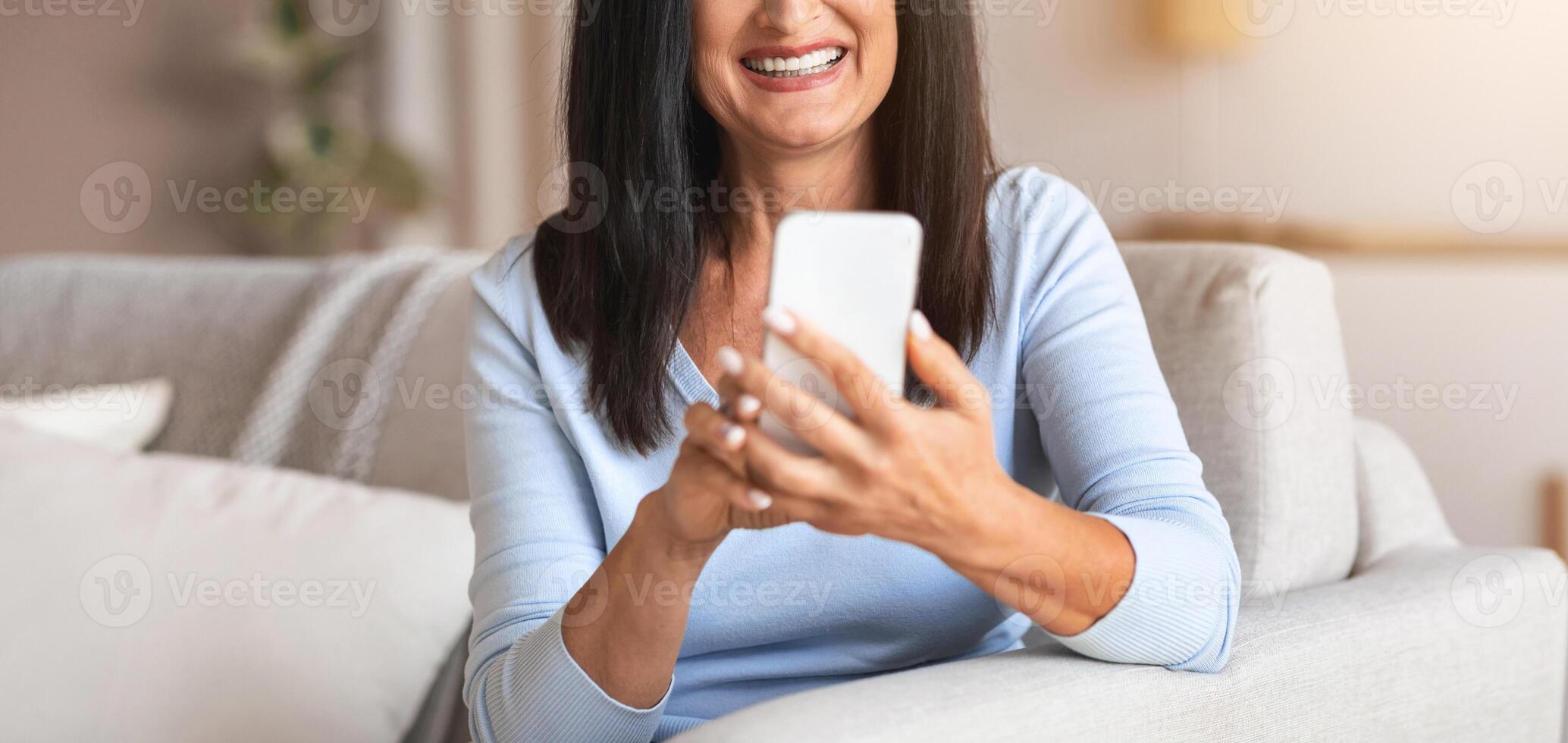 Weekend Pastime. Portrait of smiling mature lady holding smart phone, typing sms message, sitting on the couch at home. Communication Concept. Happy woman surfing internet, watching funny photo