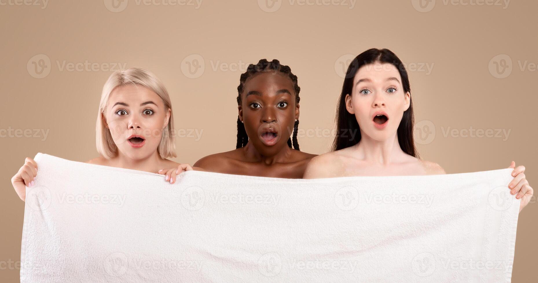 Three Emotional Multi-Ethnic Women Hiding Behind Bath Towel With Shocked Face Expression, Group Of Dazed Ladies Standing With Open Mouth Over Beige Background, Reacting To Amazing Offer, Copy Space photo