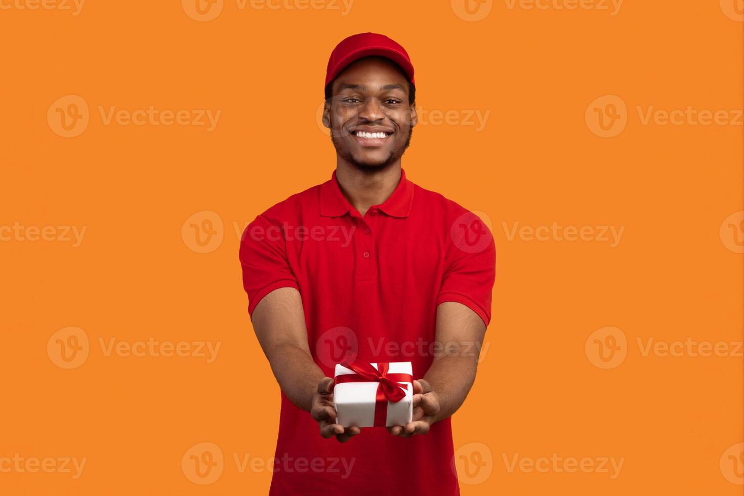 This Present Is For You. Front view portrait of smiling black delivery man holding wrapped gift box giving it to the camera, isolated on orange studio background, greeting with special day photo