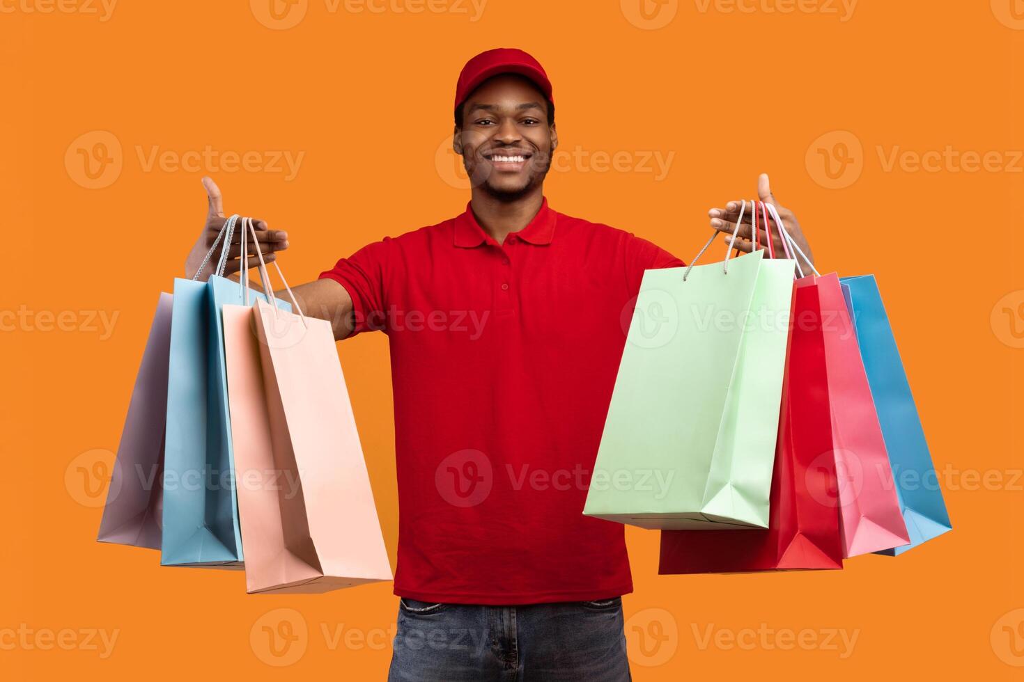 Fast Shipment Concept. Cheerful black deliveryman wearing red uniform and cap holding and showing colorful shopping bags with orders and presents, isolated over yellow orange studio background photo