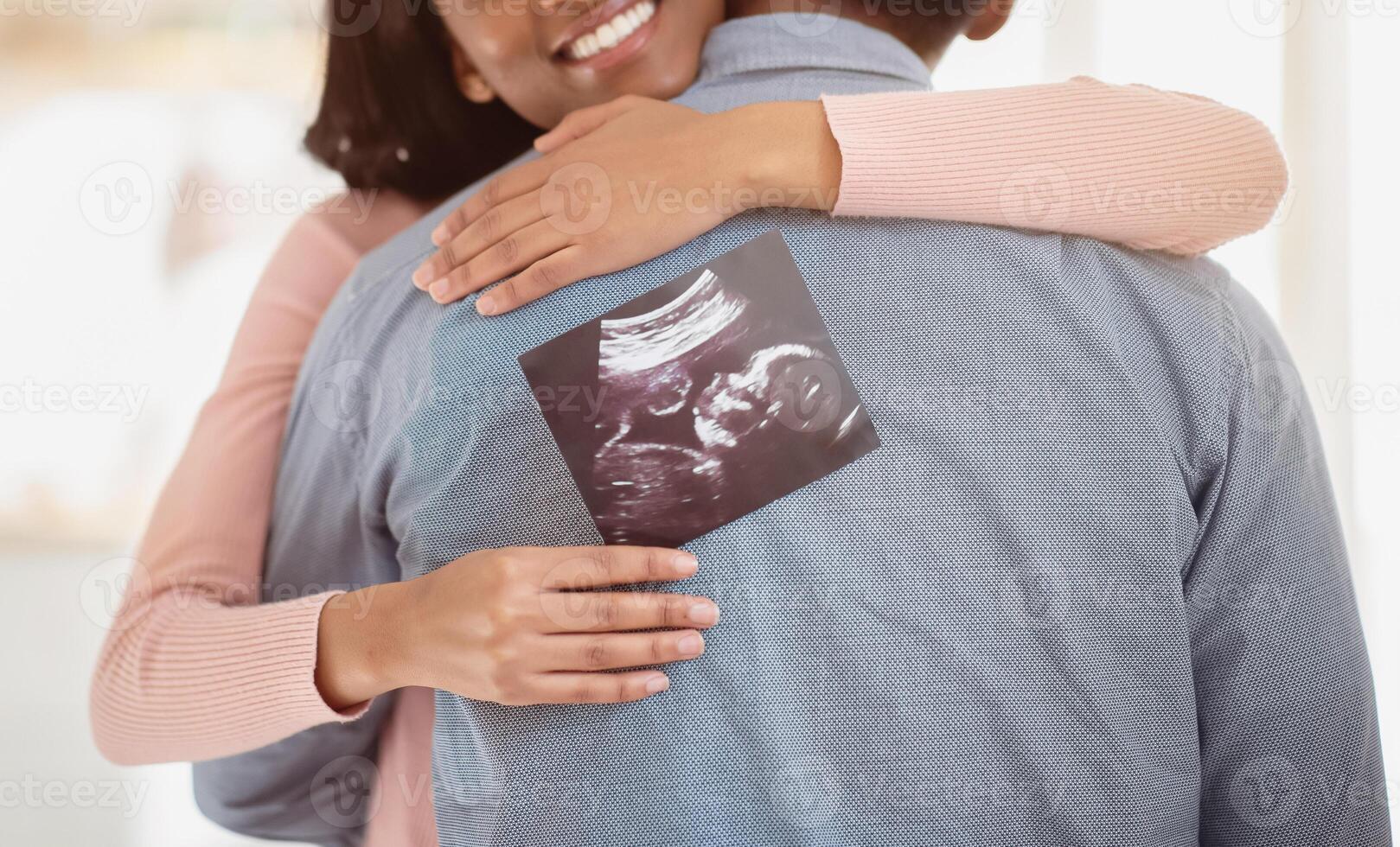 Young black pregnant woman with photo of unborn baby hugging her husband indoors, close up. Happy future mother and father with ultrasound picture of their child embracing at home