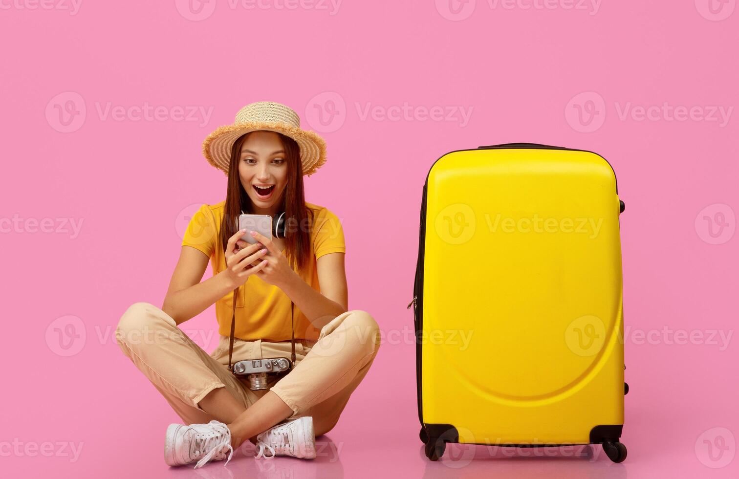 Amazed young woman sitting by suitcase and using mobile phone over pink studio background, found great deals for travelling. Active resting, vacation, tourism, travelling concept photo