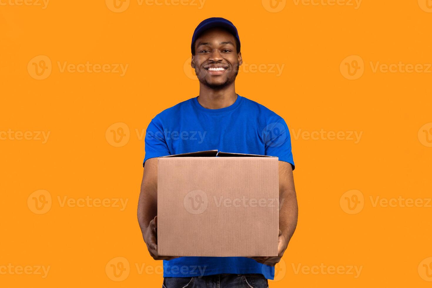 Delivery Service Concept. Smiling black mailman wearing blue cap and t-shirt holding cardboard box, showing and giving it to camera, copy space. Postal worker standing on orange studio background photo