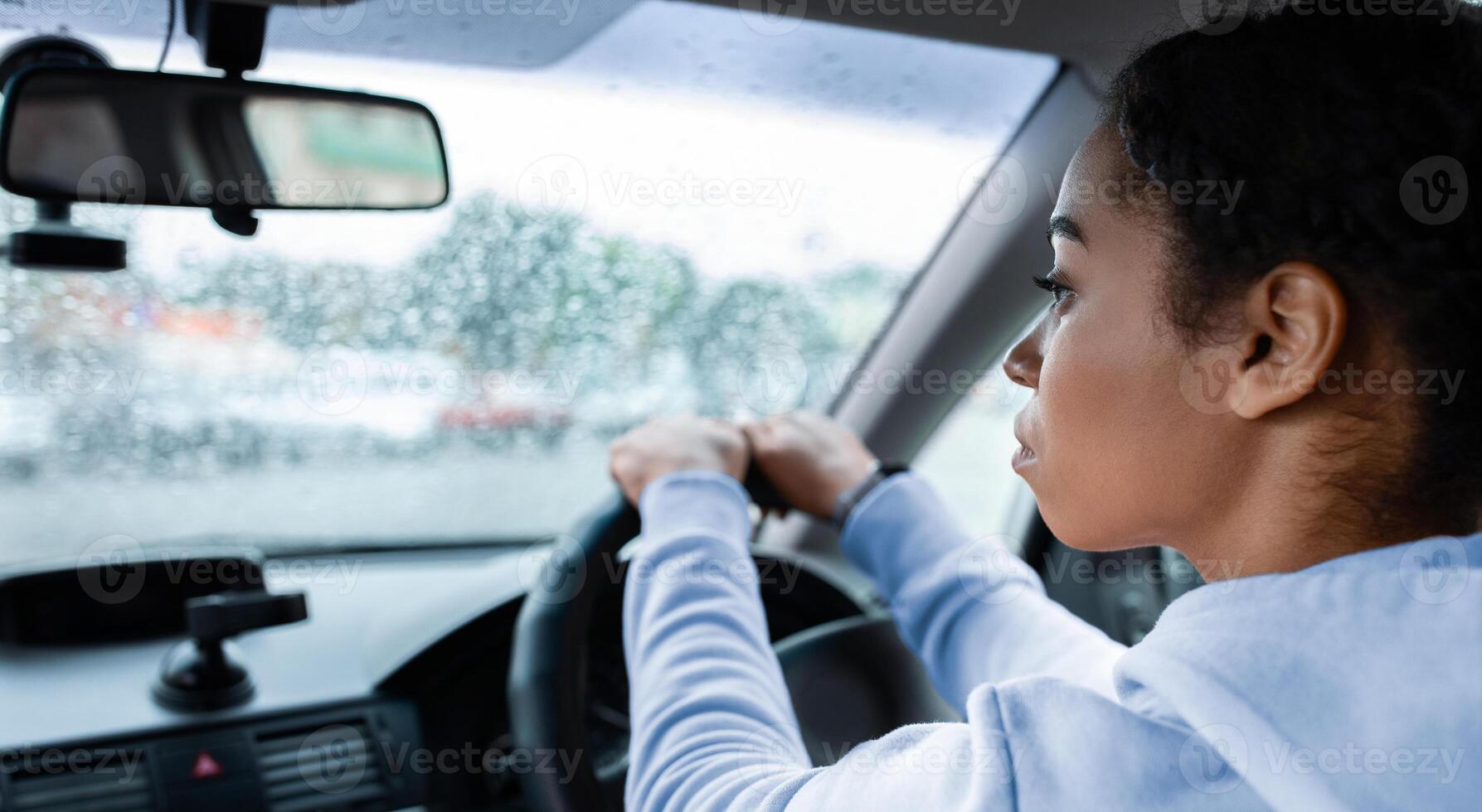 Working as taxi driver in city in bad rainy weather and way to job. Focused young african american lady drives car and holds steering wheel, looks at road on windshield with raindrops, empty space photo