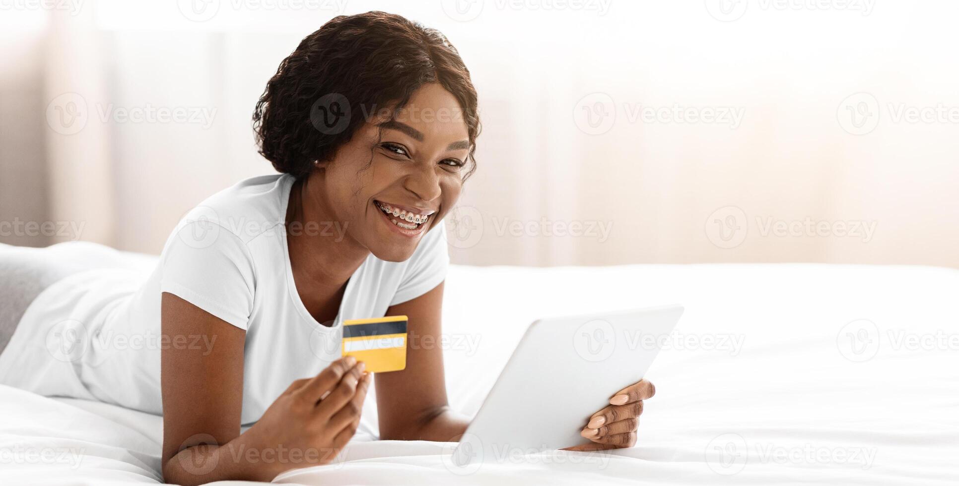Happy pretty african american woman shopping from home, using digital tablet while laying on bed, copy space. Joyful black lady holding credit card, using mobile application for e-commerce on her pad photo