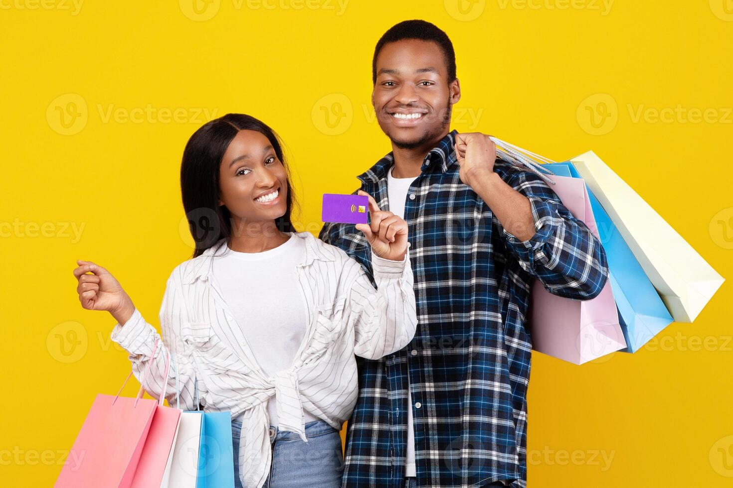 Fun shopping together and modern payment. Smiling young african american students couple with many colored shopping bags show credit card, isolated on yellow background, empty space, studio shot photo