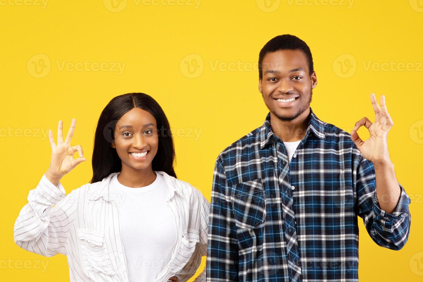 Good mood sign. Happy millennial african american man and woman in casual, show OK with hands and look at camera during covid-19 quarantine, isolation on yellow background, studio shot, copy space photo
