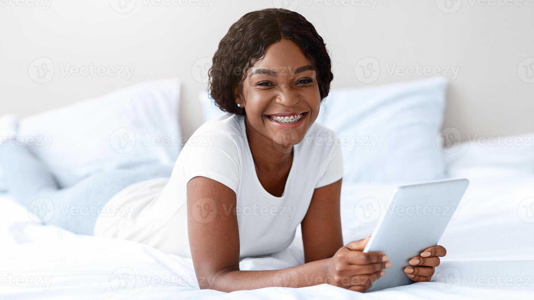 Pretty black woman with digital tablet smiling at camera, enjoying new useful applications for pad, reading book or watching something online, surfing on social media, bedroom interior, copy space photo