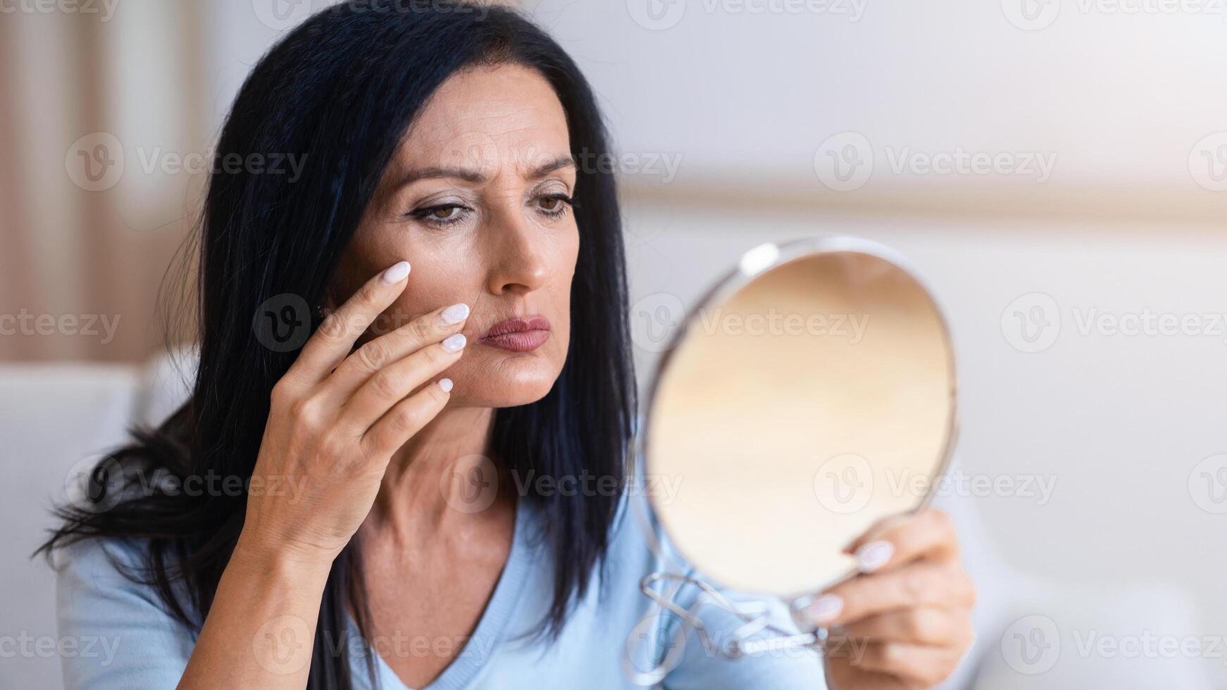 Aged Skin. Portrait of serious senior woman with long dark black hair holding mirror and looking at her reflection, cheking and examining facial wrinkles, doing morning skincare routine at home photo